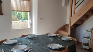 Uma sala de jantar com uma mesa redonda posta para seis pessoas, cadeiras de madeira, uma luminária pendente e uma escadaria ao fundo.