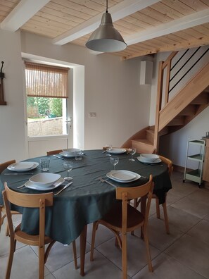 Una zona de menjador amb una taula rodona preparada per a sis, cadires de fusta, una làmpada penjant i una escala al fons.