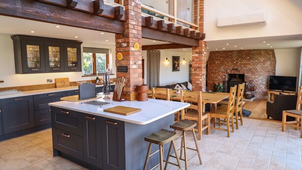 A spacious kitchen with dark cabinetry, a large island, and a brick fireplace. - Brambleberry Barn (Spilsby)