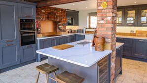 A modern kitchen with grey cabinets, a central island with stools, a brick oven, and a large window. - Brambleberry Barn (Spilsby)