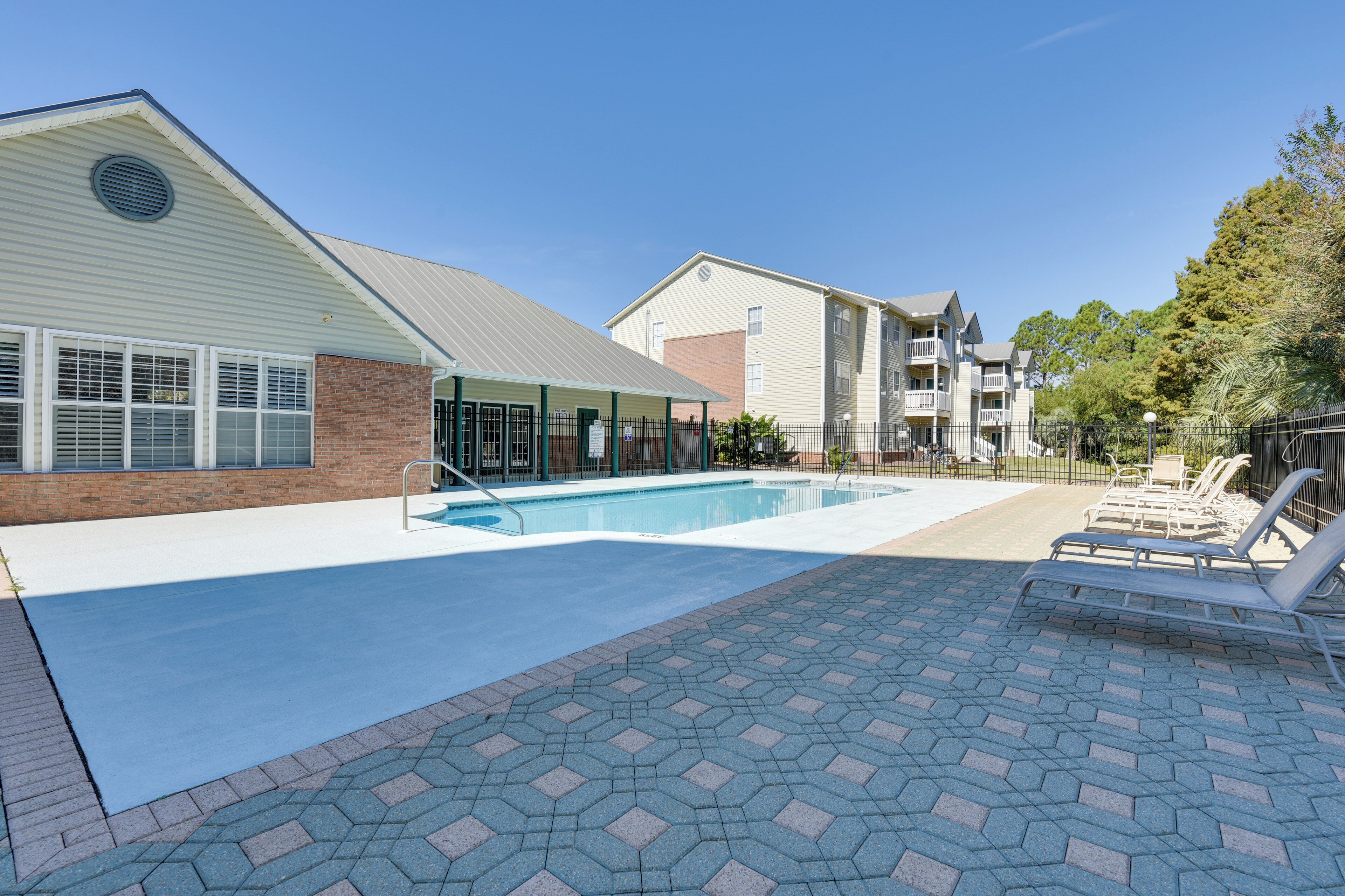 A swimming pool with a covered area and lounge chairs, surrounded by a brick walkway and buildings.