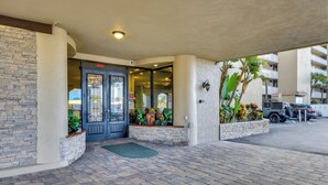 Entrada de hotel con fachada de piedra, puerta de vidrio y maceta con plantas tropicales.