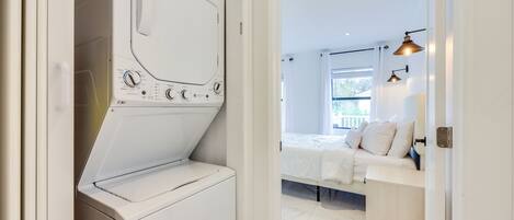 A modern laundry room with a front-loading washing machine, a bed with white bedding, and a view of a balcony through a window.