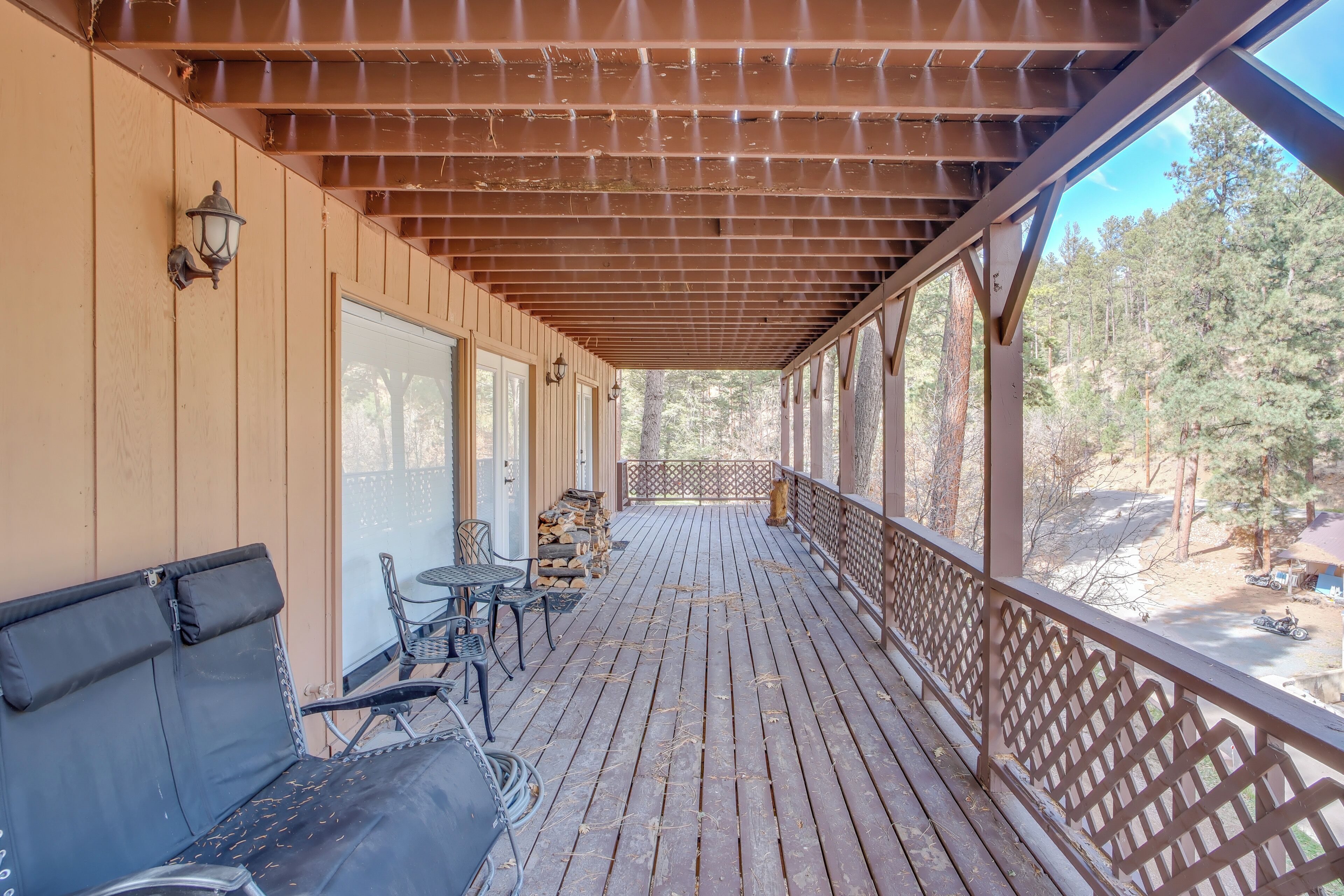 A wooden deck with a bench and chairs, a stack of firewood, and a view of a forested area.