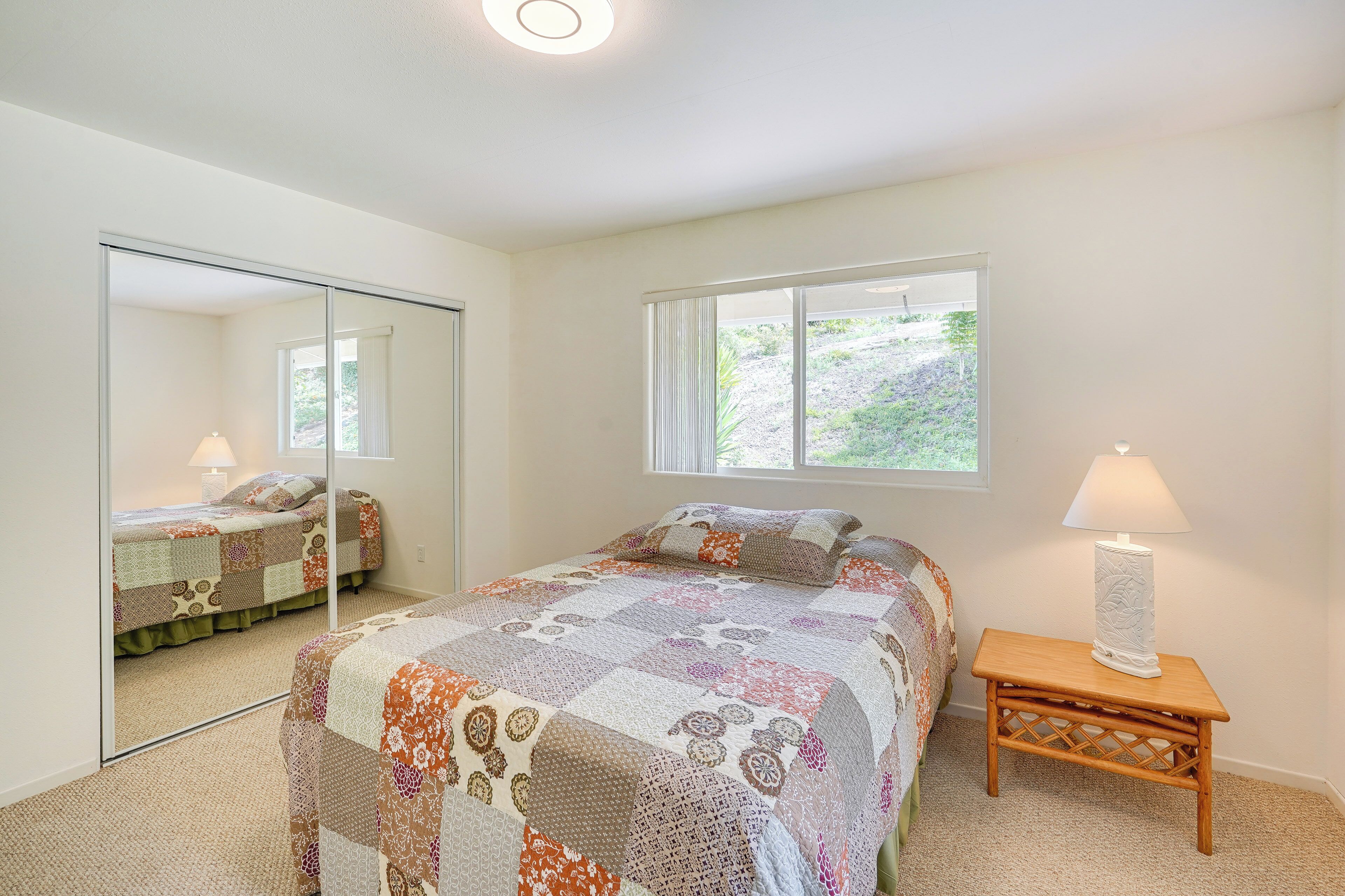 A bedroom with a large mirror, a bed with a patterned quilt, a bedside lamp, and a window with a view of greenery.