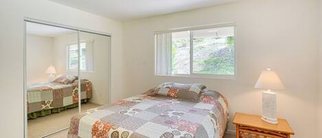 A bedroom with a large mirror, a bed with a patterned quilt, a bedside lamp, and a window with a view of greenery.
