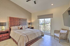 Une chambre avec un grand lit, une tête de lit en bois, deux tables de chevet avec des lampes, un fauteuil en osier et un balcon avec vue.