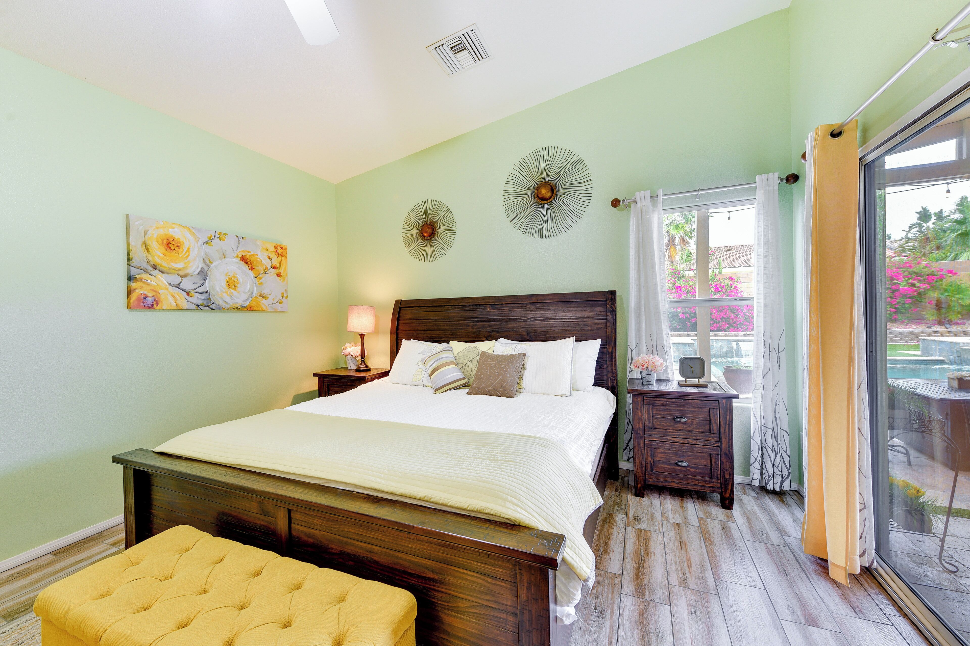 A bedroom with a large bed, a wooden nightstand, a yellow ottoman, and a painting on the wall.