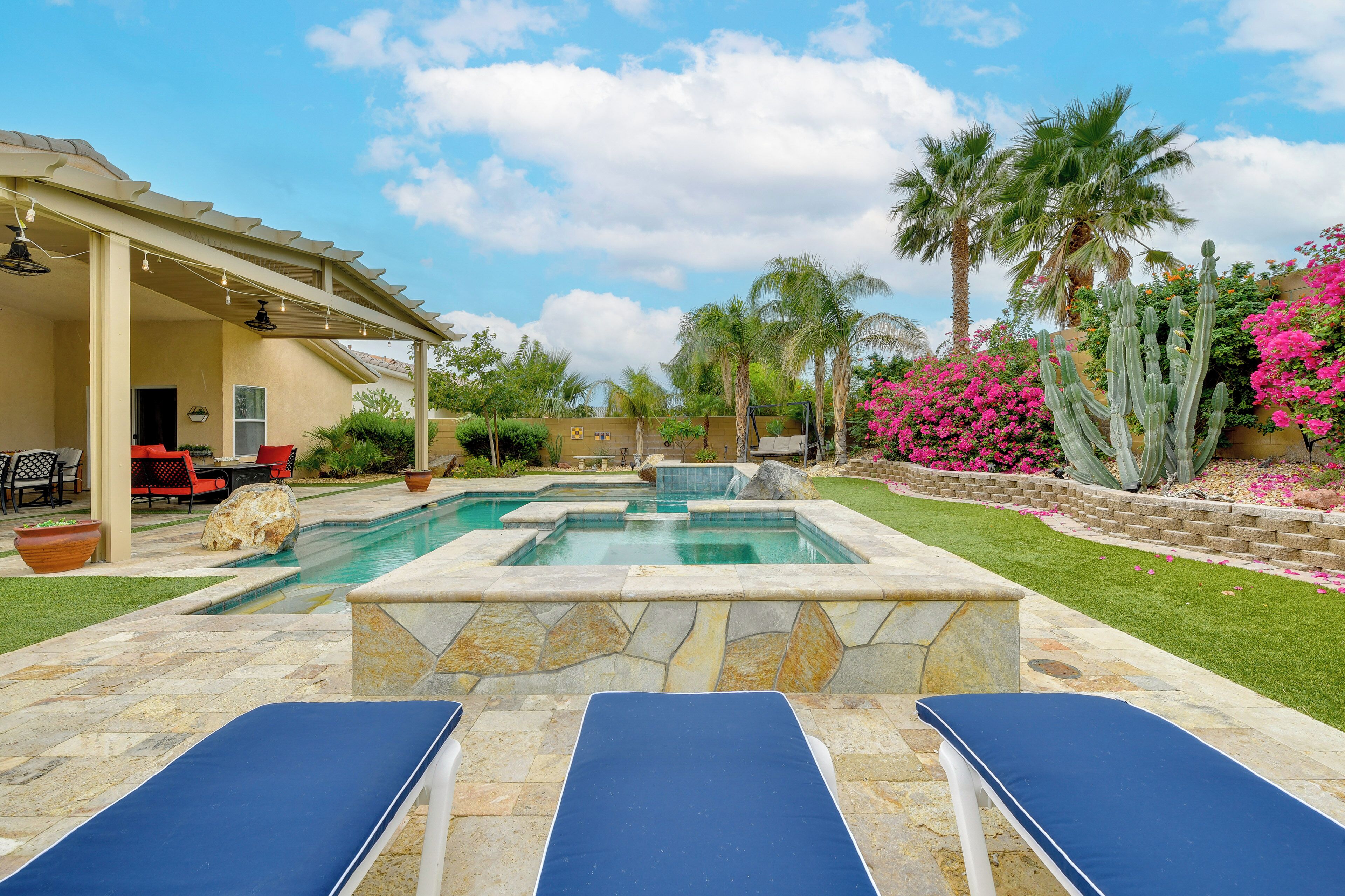 A backyard with a swimming pool, lounge chairs, a covered patio, palm trees, and flowering plants.