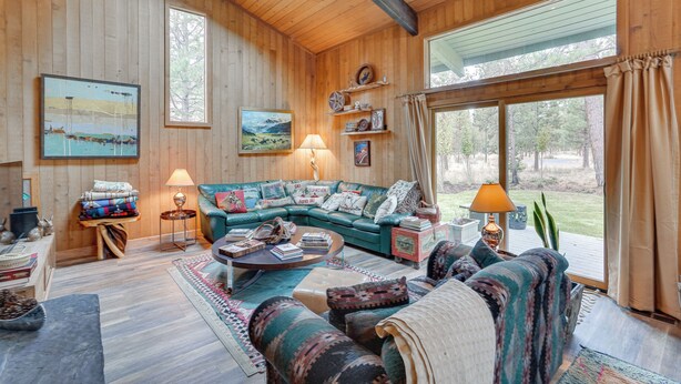A cozy cabin interior with a large sofa, a coffee table, and a fireplace. There are wooden walls and a large window offering a view of the outdoors. - Sunriver Resort Home: Near Skiing, Town & Trails (Bend)