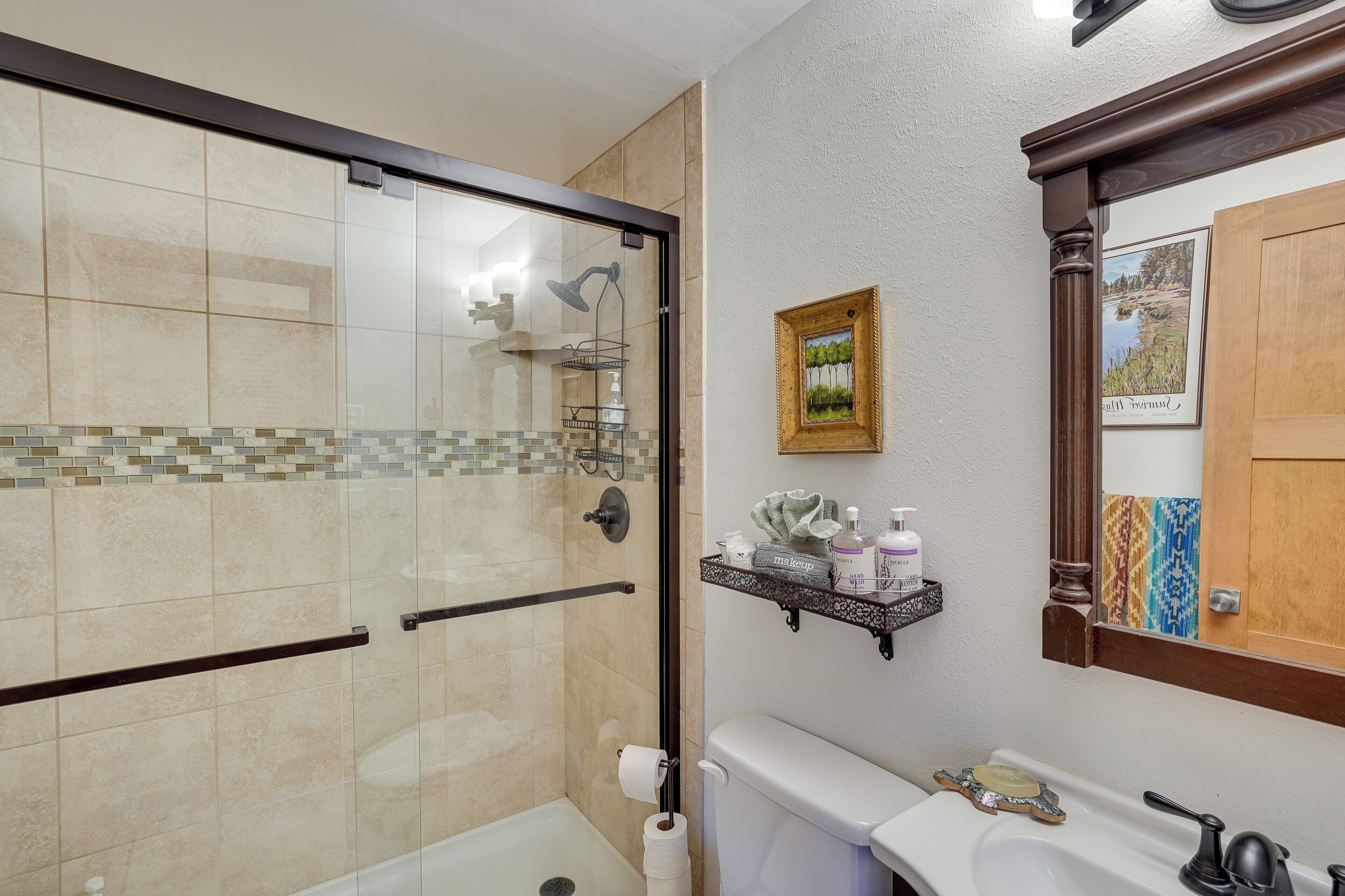 A bathroom with a glass-enclosed shower, a toilet, a sink with a soap dish, and a shelf with toiletries.