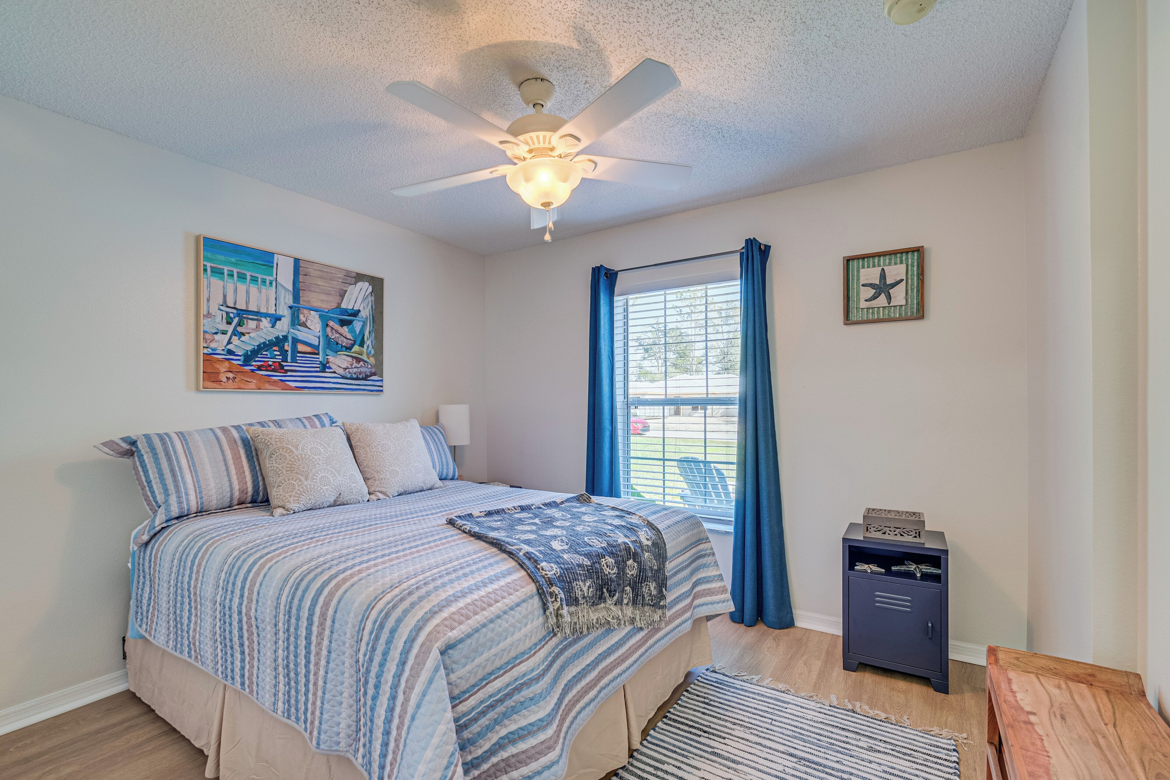 A bedroom with a bed, a ceiling fan, a window with curtains, a nightstand, and a painting on the wall.