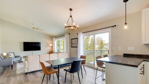 A modern living room with a sofa, dining table, and chairs, a kitchen area with a countertop, and a large window providing a view of the outdoors.