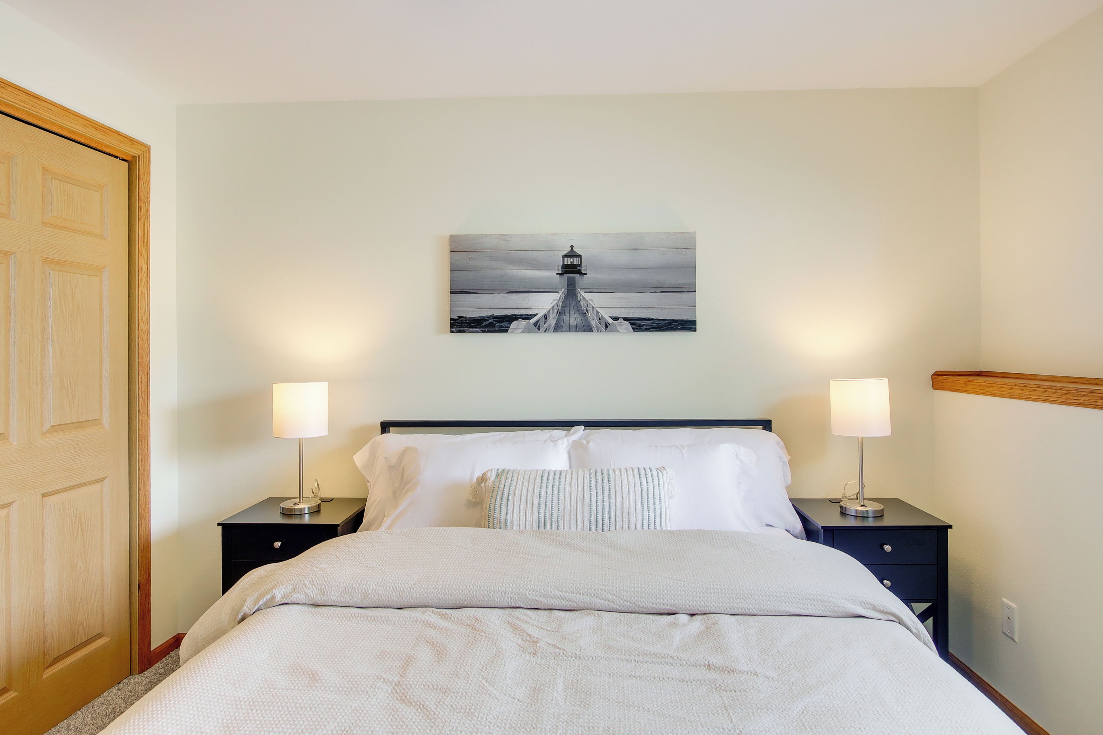 A neatly made bed with white linens, two bedside tables with lamps, and a painting of a lighthouse on the wall.