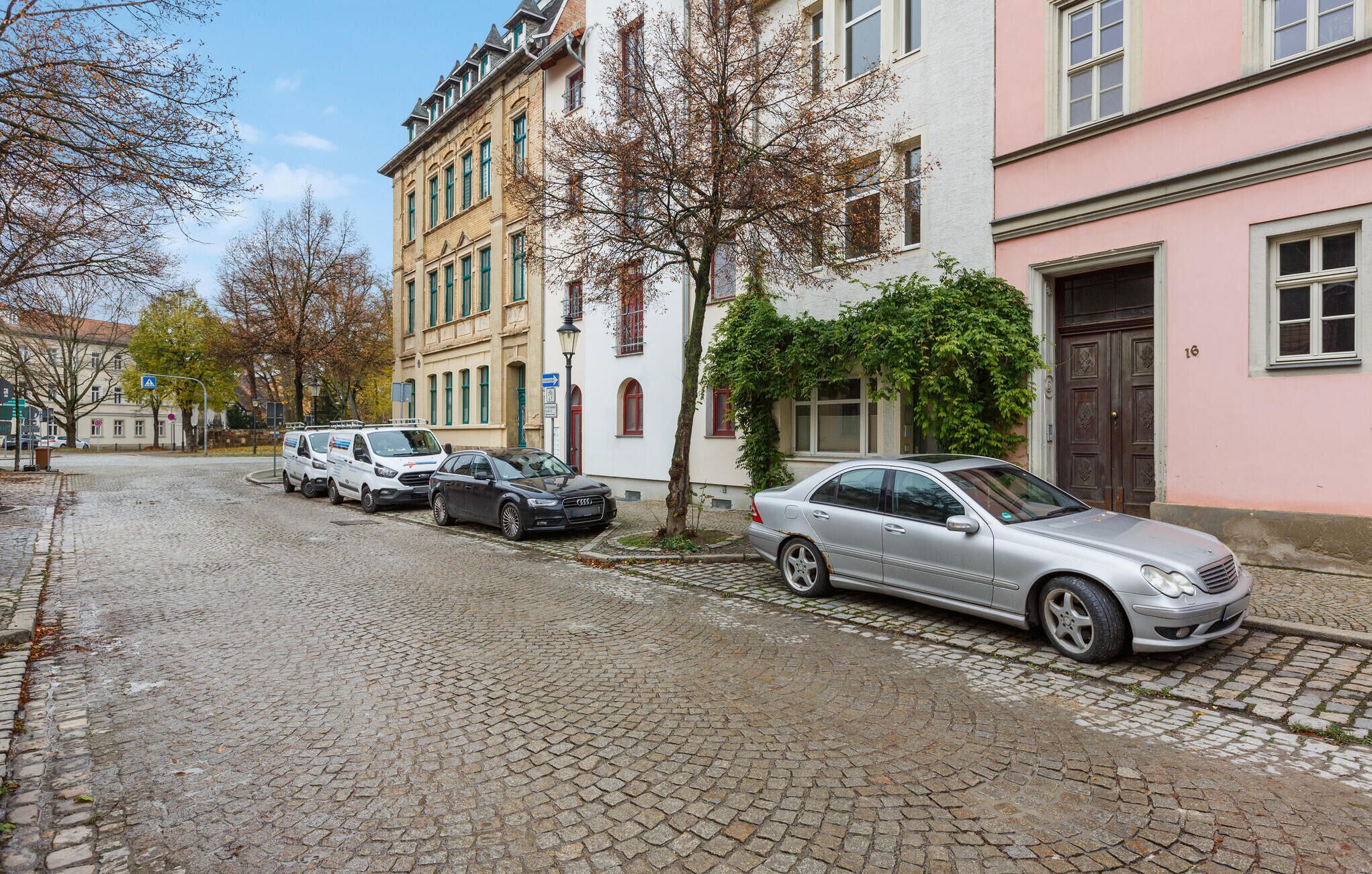 Une rue pavée bordée de bâtiments historiques, de voitures garées et d’un panneau indiquant un passage piéton.