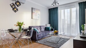 A modern living room with a glass dining table, clear chairs, a purple sofa, and a large window with curtains. - Mokotów Domaniewska 47a (Warsaw)