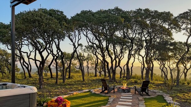 A patio with a hot tub, two chairs, and a fire pit surrounded by a stone pathway and lush greenery. - Splendor in the Hills, Wimberley, Putting Green, Views, Hot tub! (Wimberley)