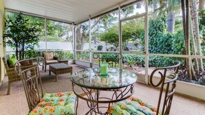 Une terrasse couverte, aménagée avec des meubles en osier, une table en verre et offrant une vue sur une végétation luxuriante.