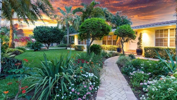 Un jardin bien entretenu, agrémenté de diverses plantes et fleurs, un chemin pavé et un bâtiment doté de grandes fenêtres et d’un toit en tuiles.