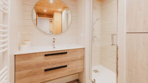 Une salle de bain moderne avec un meuble-lavabo en bois, un miroir rond, une cabine de douche et un mur recouvert de carreaux blancs.