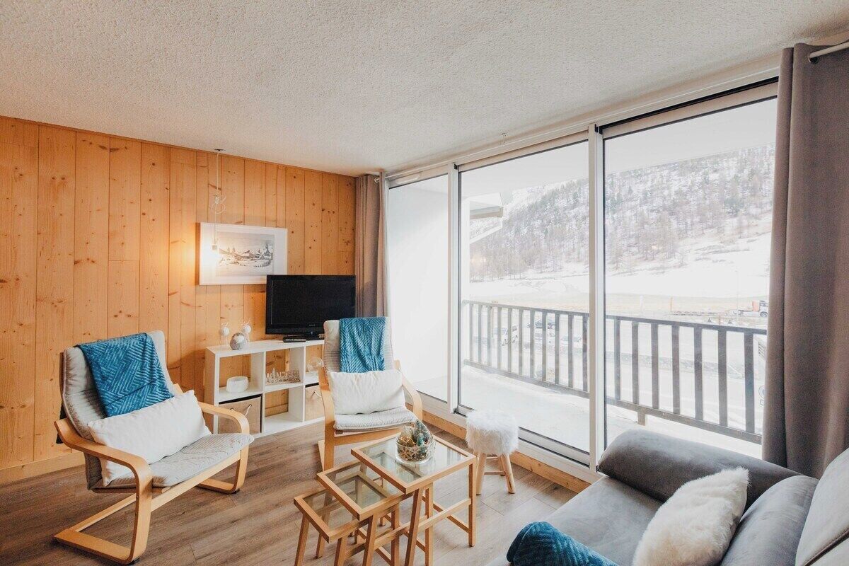 Un salon moderne avec des murs en bois, un téléviseur à écran plat, un canapé, deux fauteuils à bascule, une petite table et un balcon offrant une vue sur un paysage enneigé.