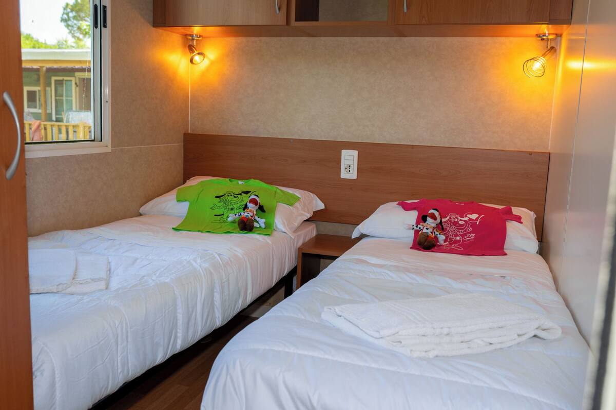 Dos camas individuales con ropa blanca, cada una con una almohada y una manta. Hay luces fijadas a la pared sobre las camas.
