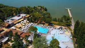 Eine Luftaufnahme eines Resorts mit einem großen Schwimmbad, umgeben von Liegestühlen und Sonnenschirmen, einer anliegenden Anlegestelle am See und verschiedenen Gebäuden mit unterschiedlichen Dachfarben.