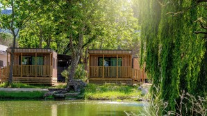 Zwei Holzhütten an einem ruhigen See mit einer Holzterrasse und einem Kiesweg, der zu ihnen führt.