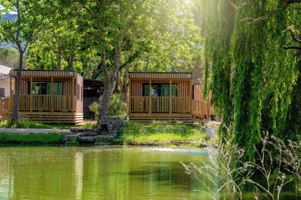 Dos cabañas de madera junto a un lago tranquilo, con un muelle de madera y un sendero de piedra que las lleva.