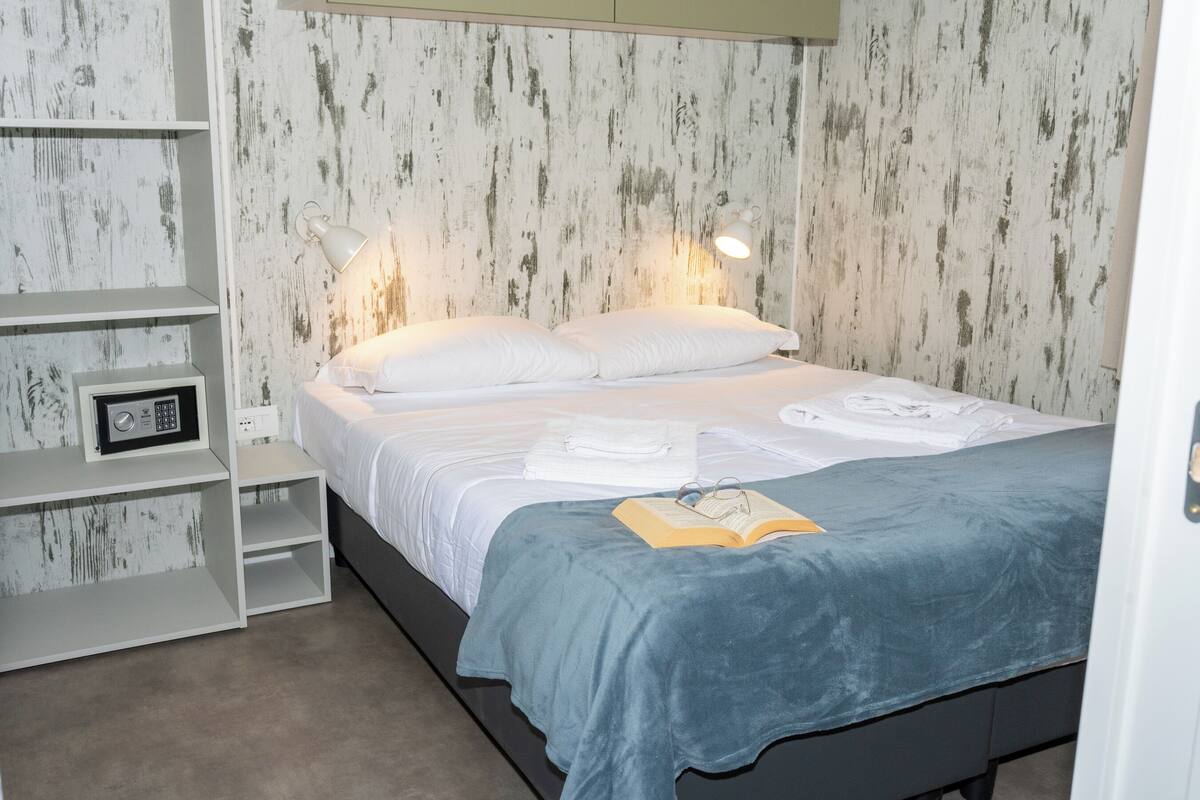 Una cama bien hecha con una colcha azul, almohadas blancas y un libro encima. Hay dos mesitas de noche con lámparas y una pequeña estantería con una radio. Las paredes tienen un diseño de paneles de madera desgastada.