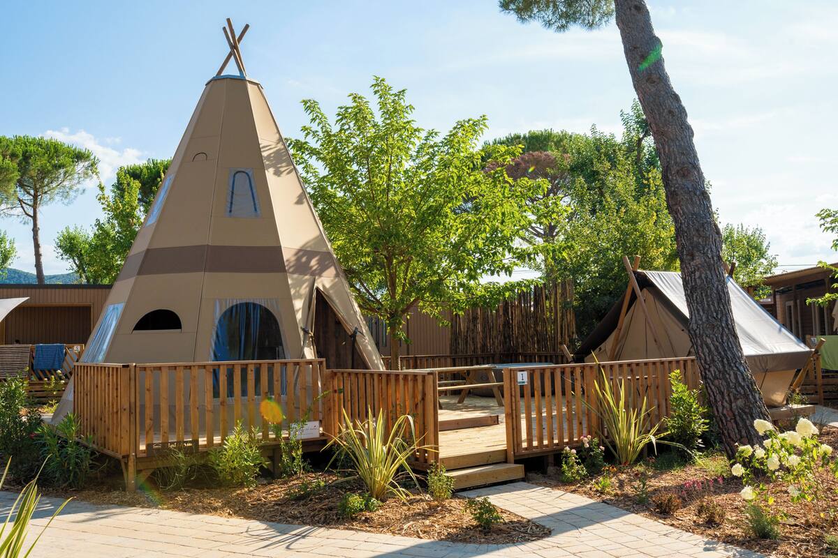 Estructura de tienda cónica con plataforma de madera y una ventana pequeña, rodeada por una valla de madera y vegetación.