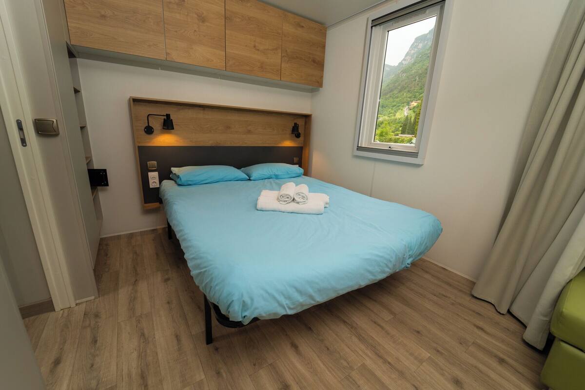 Un dormitorio con cama, cabecero de madera, lámparas de noche y ventana con vista a follaje.