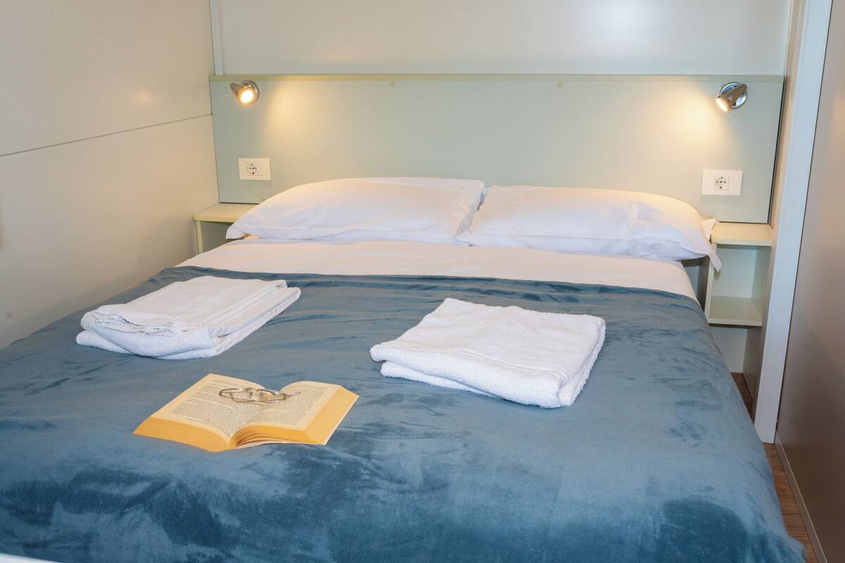 Una cama bien hecha con una colcha azul, dos almohadas blancas y dos toallas dobladas. Un libro abierto está sobre la cama.