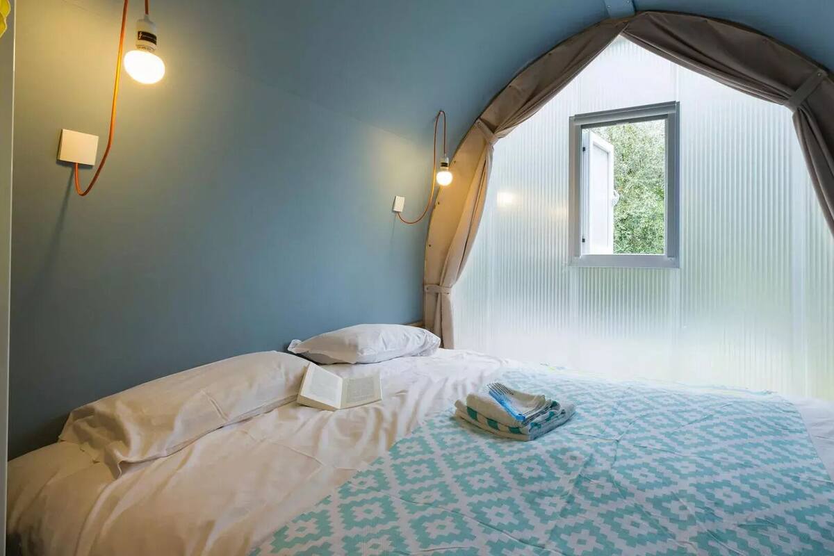 Un dormitorio con cama, almohadas, mesita de noche, ventana con cortinas e iluminación en la pared.