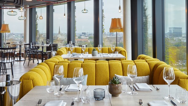 Een restaurant met geel gestoffeerde zitplaatsen, grote ramen en een uitzicht over de stad.