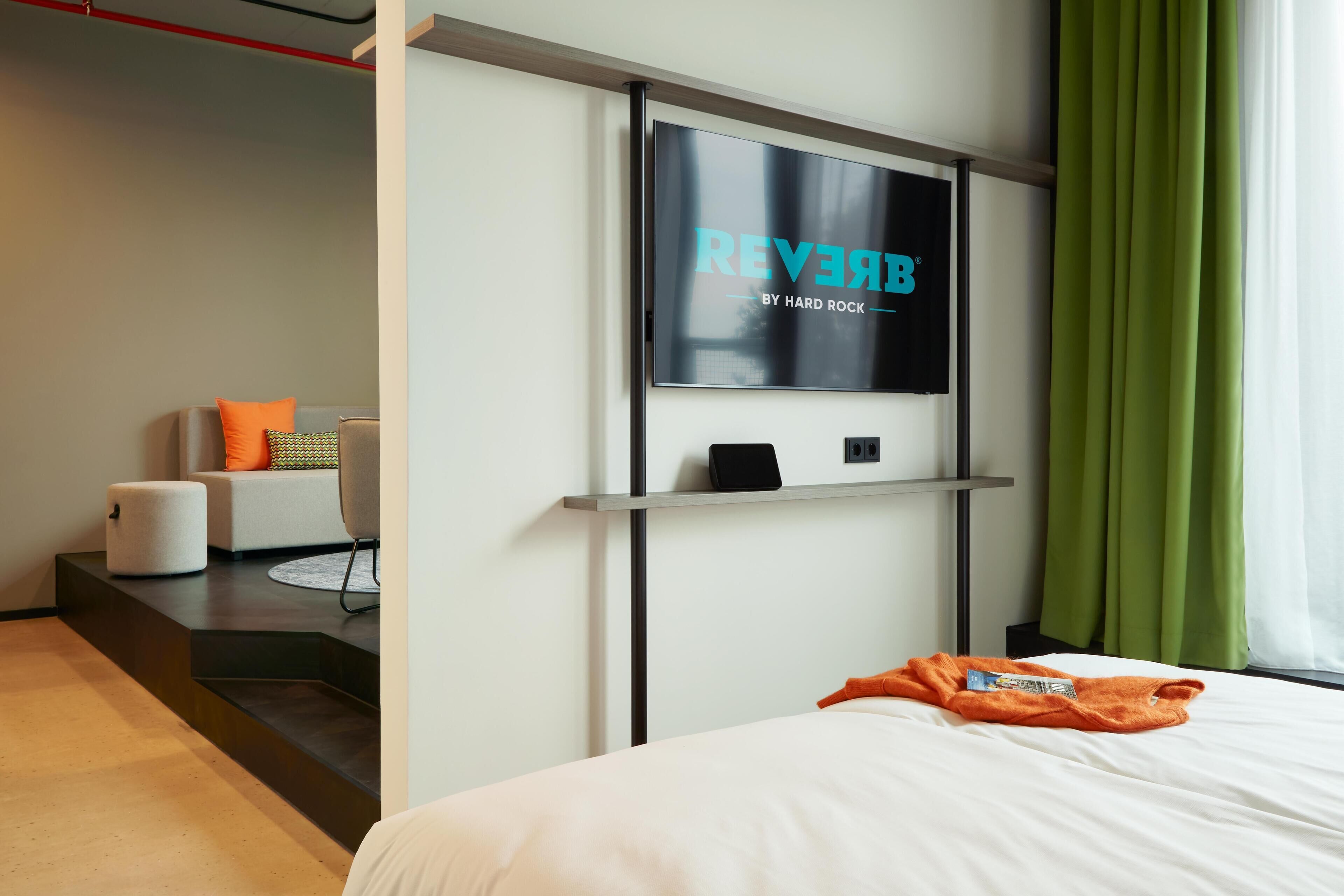 A modern hotel room with a large flat-screen TV mounted on the wall, a bed with a white comforter, a small seating area with a chair and a sofa, and a green curtain on the window.