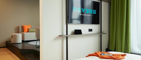 A modern hotel room with a large flat-screen TV mounted on the wall, a bed with a white comforter, a small seating area with a chair and a sofa, and a green curtain on the window.