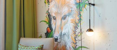 Habitación con un mural de un zorro, un sofá con un cojín estampado, una mesita y una lámpara colgante.