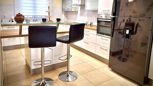 Una cocina moderna con un refrigerador de acero inoxidable, horno empotrado e isla de vidrio con dos taburetes negros.