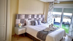 Un dormitorio con una cama grande, una mesita de noche, un ventilador de techo y vistas a un paisaje verde por la ventana.