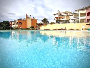 En swimmingpool med klart, blåt vand, omgivet af bygninger med balkoner og et palmetræ.