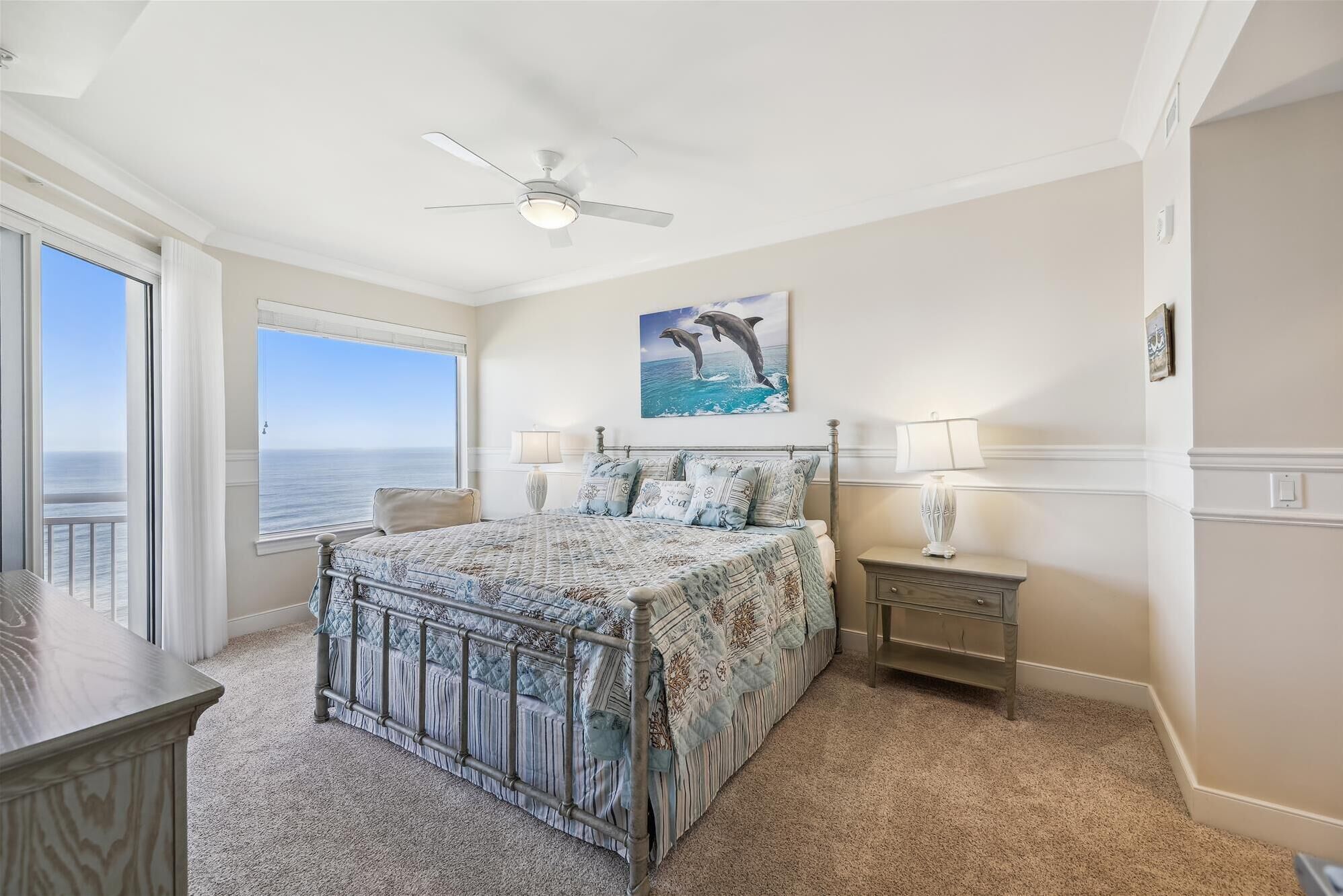 Une chambre à coucher avec un lit, un ventilateur de plafond, une table de chevet, une lampe, un tableau représentant des dauphins et une vue sur l’océan par la fenêtre.