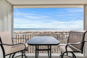 Un balcone con due sedie e un tavolo, affacciato su una spiaggia con una recinzione e un cielo sereno.