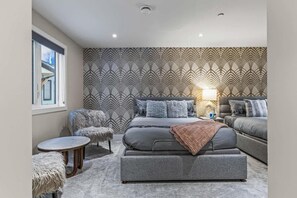 Una camera da letto moderna con un letto grande, un divano e due pouf. Si nota una finestra sul lato sinistro e una parete decorata dietro il letto.