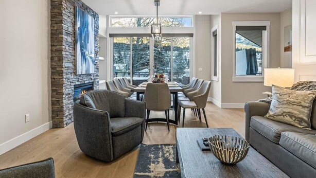 A modern living room with a fireplace, a dining area with a long table and chairs, and a sofa with a lamp. - NEW Schloss by SSR Top View HOT TUB 5BR ( BL# RES-11684 ) (Canmore)
