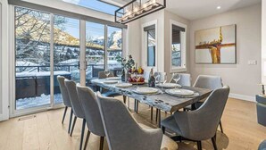 A dining area with a large table set for six, large windows offering a view of a snowy landscape, and a modern chandelier. - NEW Schloss by SSR Top View HOT TUB 5BR ( BL# RES-11684 ) (Canmore)