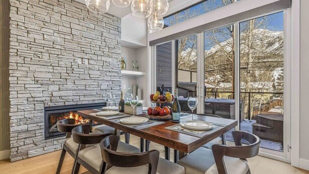 A dining area with a stone fireplace, a wooden table set for four, and a view of a snowy landscape through large windows. - NEW！ Chambord by SSR Top View HOT TUB 5BR ( BL# RES-11745 ) (Canmore)