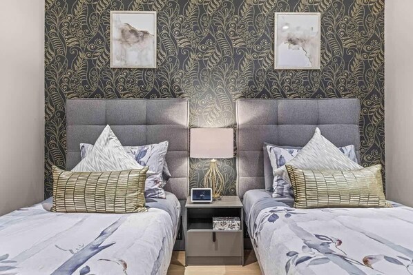 Habitación con dos camas, cada una con cabecero gris y ropa de cama con estampado floral. Hay dos mesitas de noche con lámparas y un pequeño reloj. Las paredes tienen un papel tapiz oscuro con motivos vegetales.