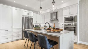 A modern kitchen with white cabinetry, a central island with bar stools, stainless steel appliances, and pendant lighting. - NEW！ Chambord by SSR Top View HOT TUB 5BR ( BL# RES-11745 ) (Canmore)