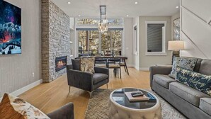 A spacious living room with a stone fireplace, a dining area, and a large window offering a view of trees. - NEW！ Chambord by SSR Top View HOT TUB 5BR ( BL# RES-11745 ) (Canmore)